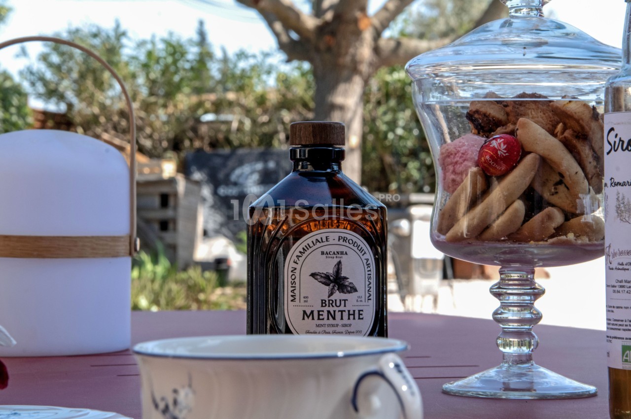 Bouteille de sirop de menthe posée sur une table avec des biscuits dans une bonbonnière et un fond extérieur ensoleillé.