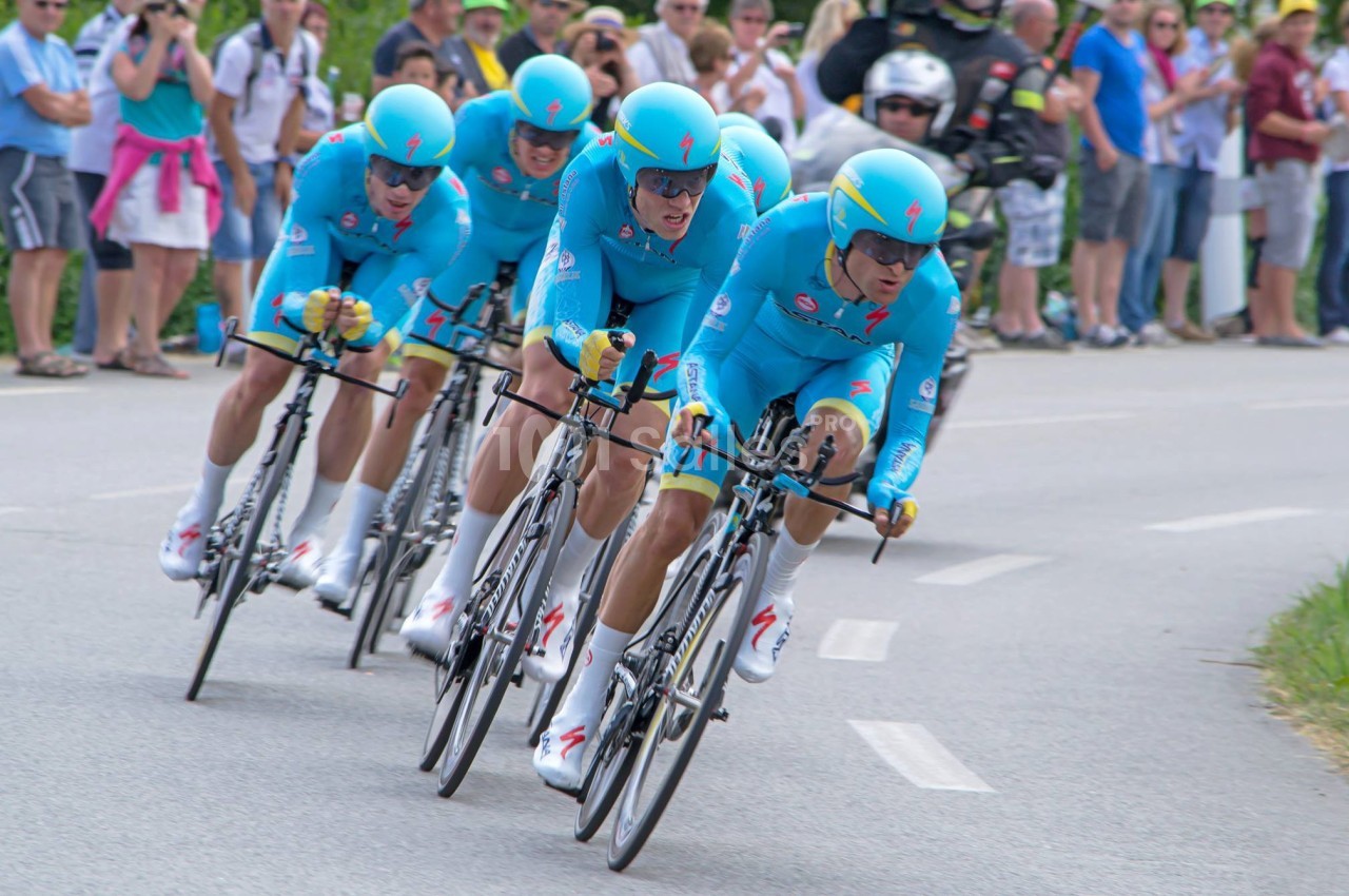 Quatre cyclistes en tenue bleue participent à une course en contre-la-montre, entourés de spectateurs au bord de la route.