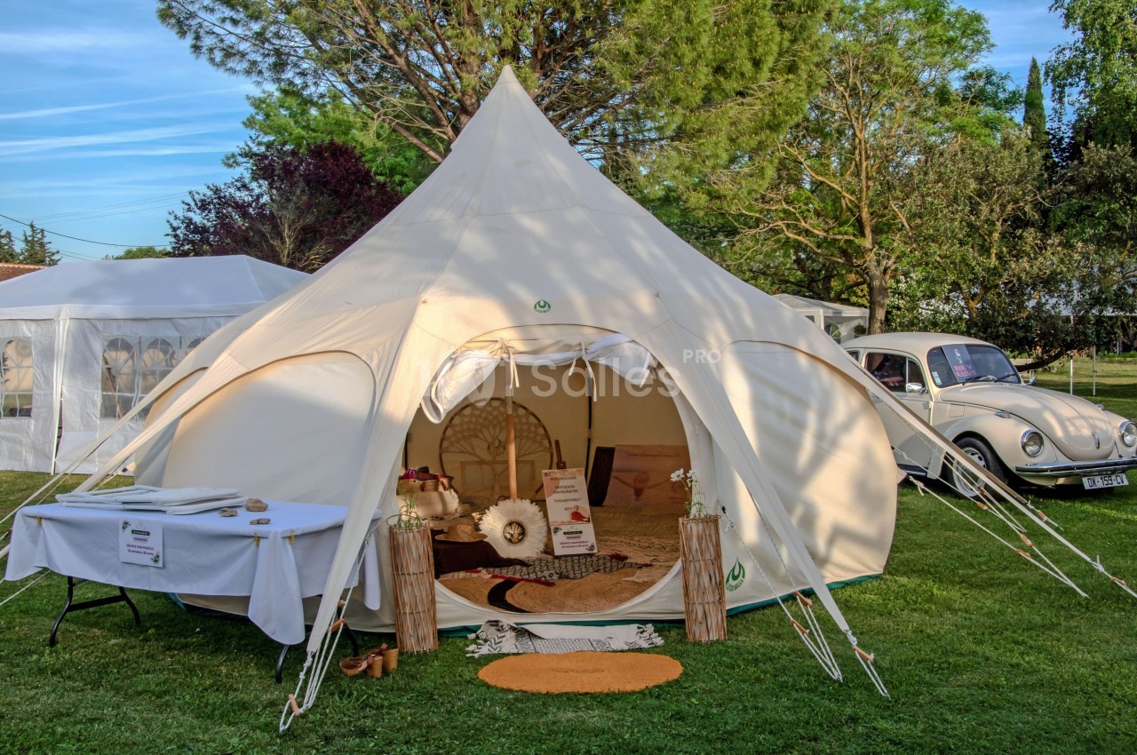 Tente beige aménagée avec des décorations bohèmes, installée sur une pelouse près d'une voiture vintage et d'arbres.