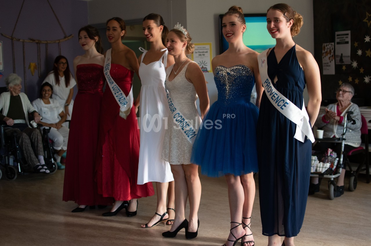 Six femmes en robes élégantes, certaines portant des écharpes de concours, posent dans une salle avec des spectateurs assis.