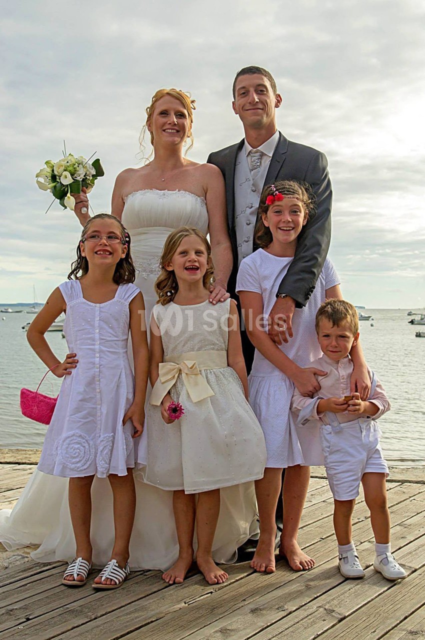 Un couple en tenue de mariage pose sur un ponton avec quatre enfants souriants devant un paysage côtier.