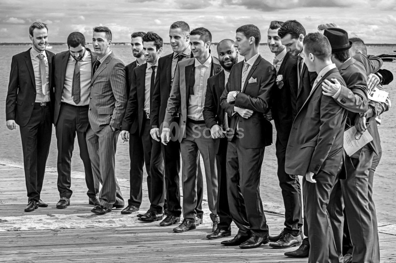 Un groupe d'hommes en costume posant sur un pont en bois près d'un plan d'eau sous un ciel nuageux.