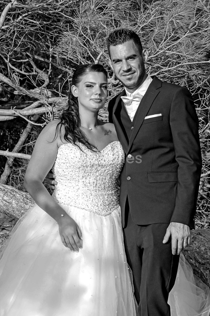 Un couple en tenue de mariage pose devant un arbre dans un cadre naturel, en noir et blanc.