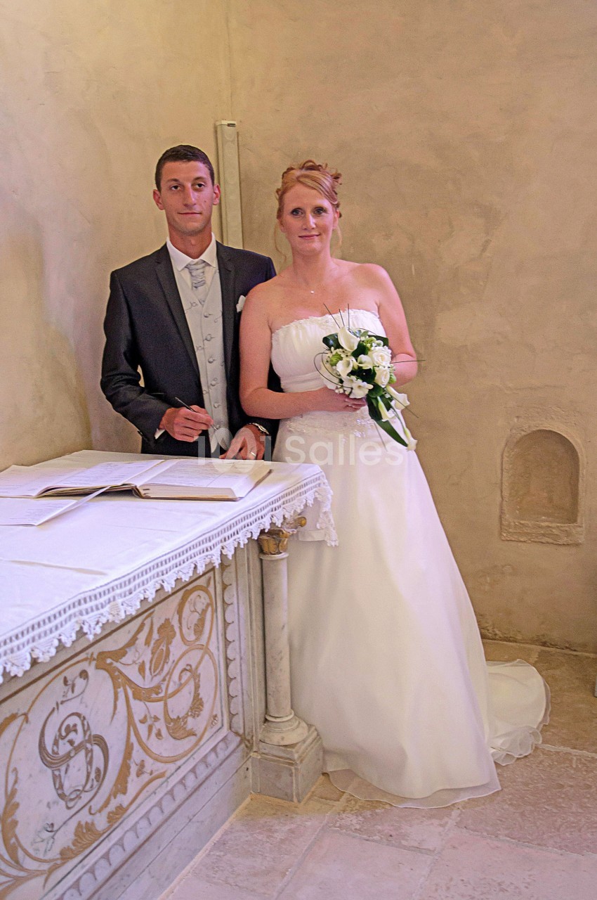 Un couple en tenue de mariage pose devant un autel décoré dans une pièce aux murs clairs.