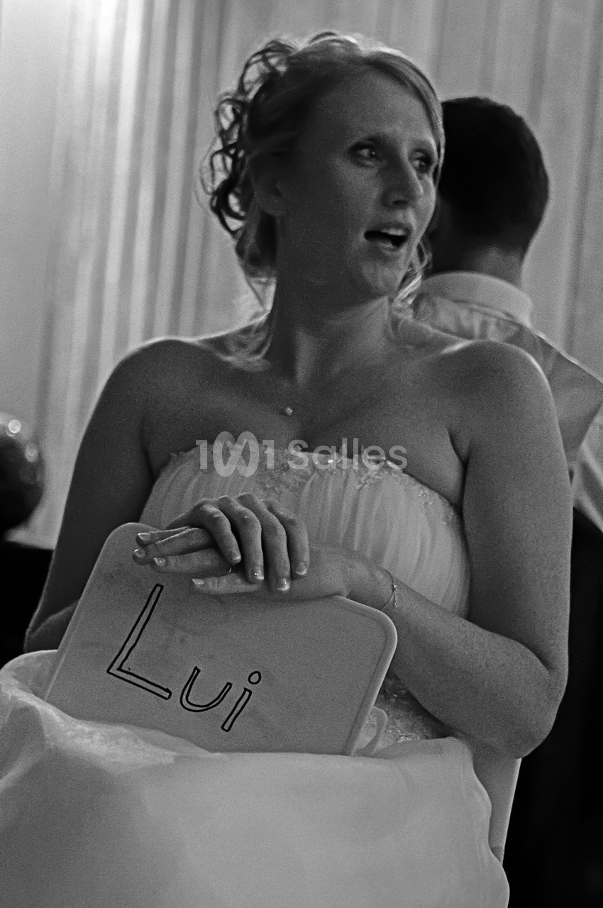 Une femme en robe de mariée tient une pancarte avec le mot ’Lui’ dans un cadre intérieur.