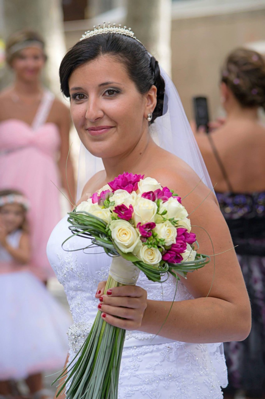 Une mariée en robe blanche tient un bouquet de fleurs blanches et roses lors d'une cérémonie en extérieur.