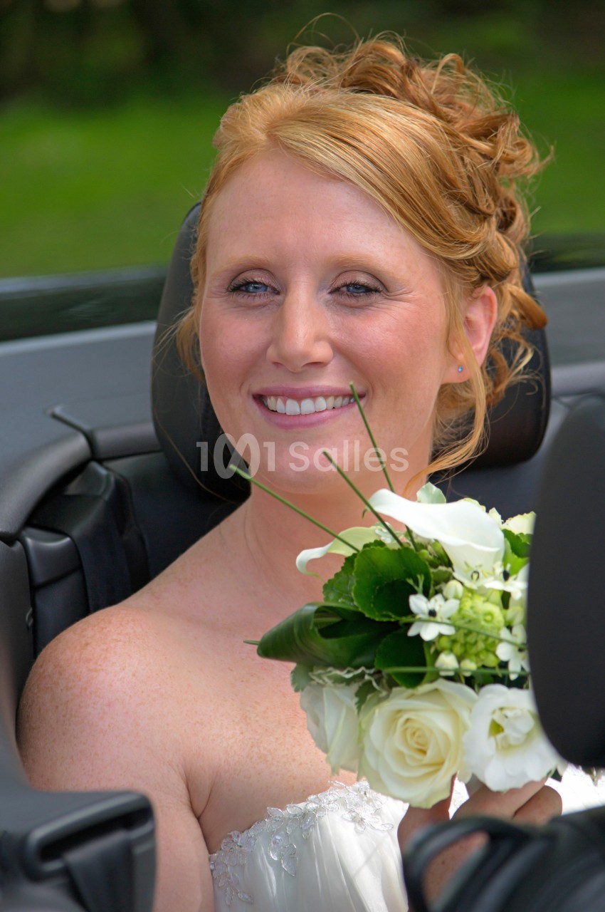 Femme souriante en robe de mariée tenant un bouquet de fleurs blanches, assise dans une voiture décapotable.