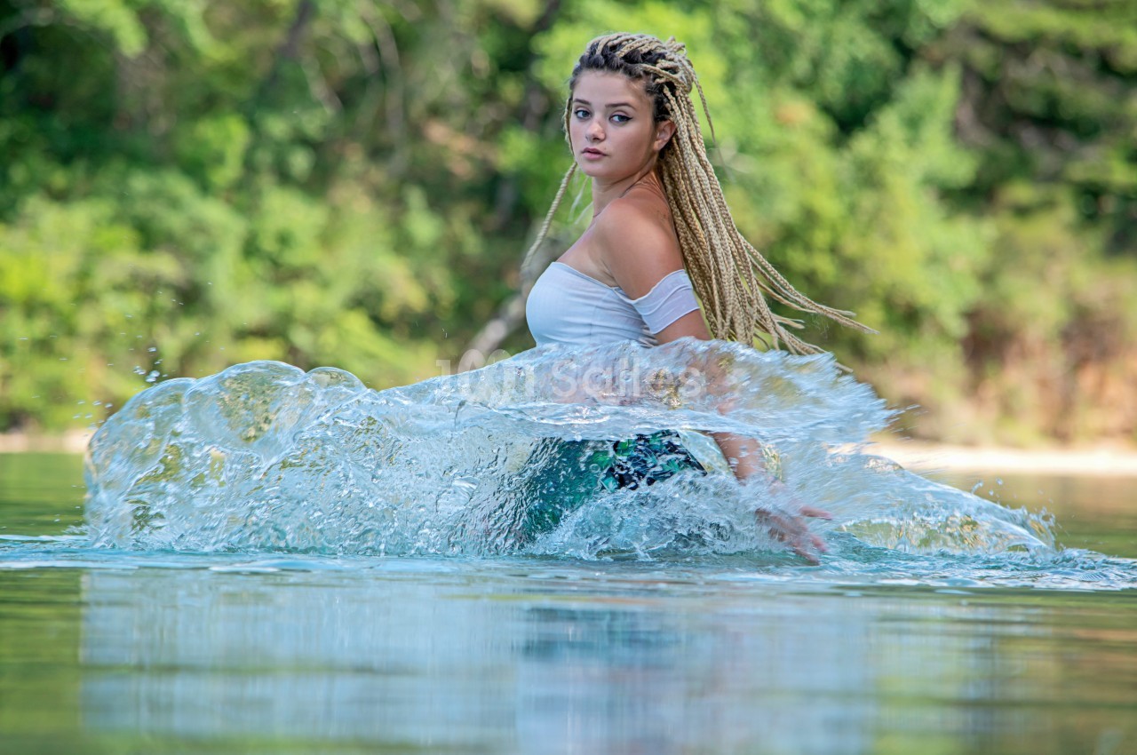Femme aux cheveux tressés debout dans l'eau, entourée d'éclaboussures, avec une forêt en arrière-plan.