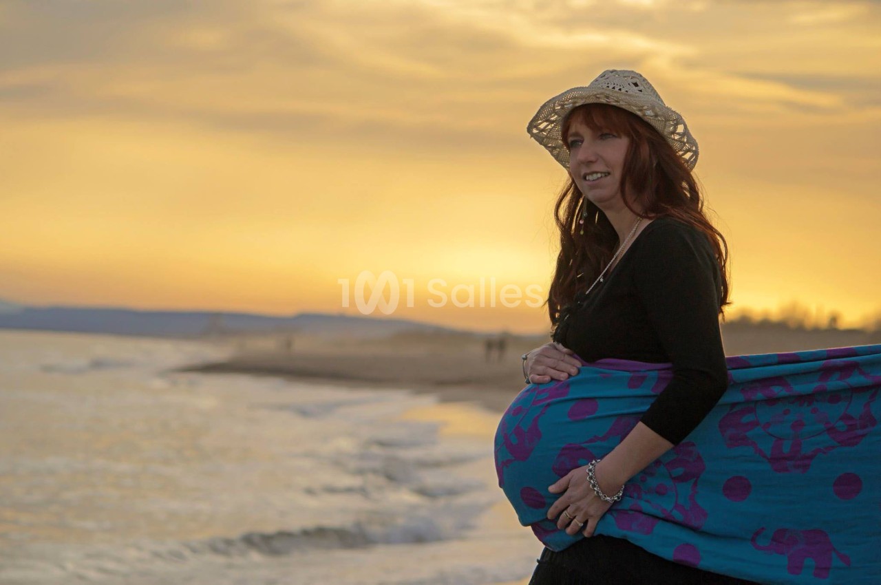 Femme enceinte souriante portant un chapeau et un tissu coloré, debout sur une plage au coucher du soleil.