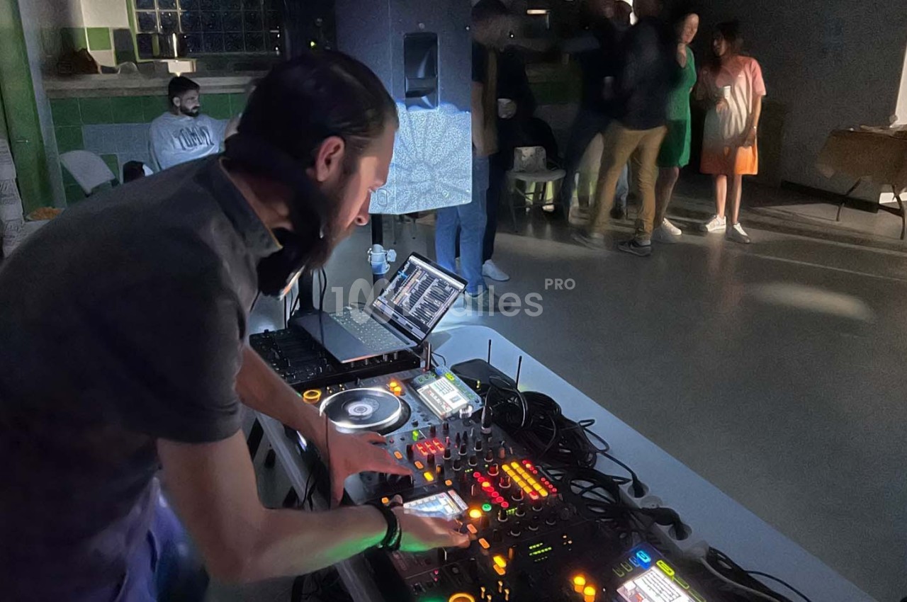 Un DJ mixe de la musique sur une console éclairée, dans une salle avec des personnes en arrière-plan.