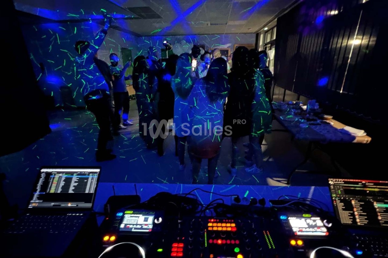 Des personnes dansent sous des lumières colorées dans une salle sombre, vue depuis une table de mixage avec équipements DJ.