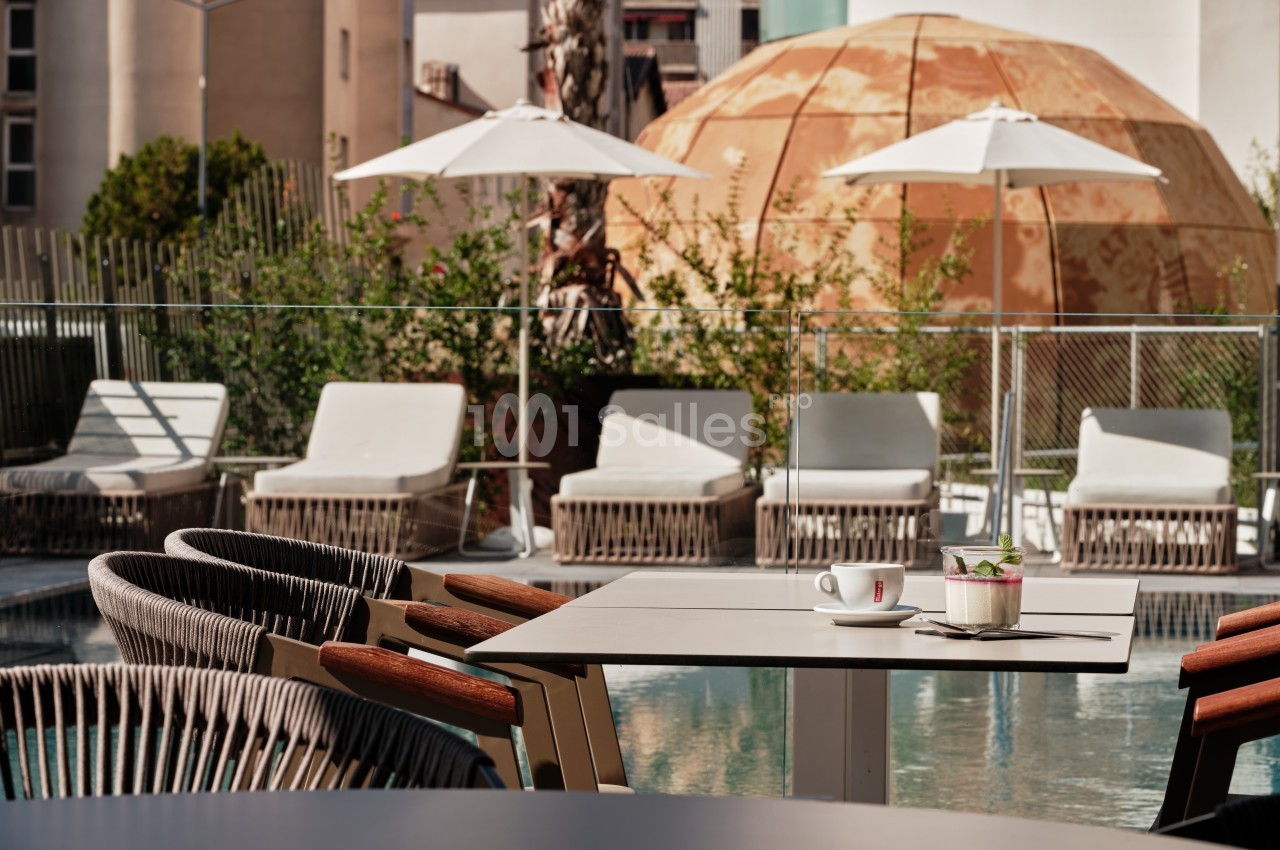 Terrasse avec tables, chaises, parasols, transats et piscine, entourée de végétation et d'un bâtiment en arrière-plan.