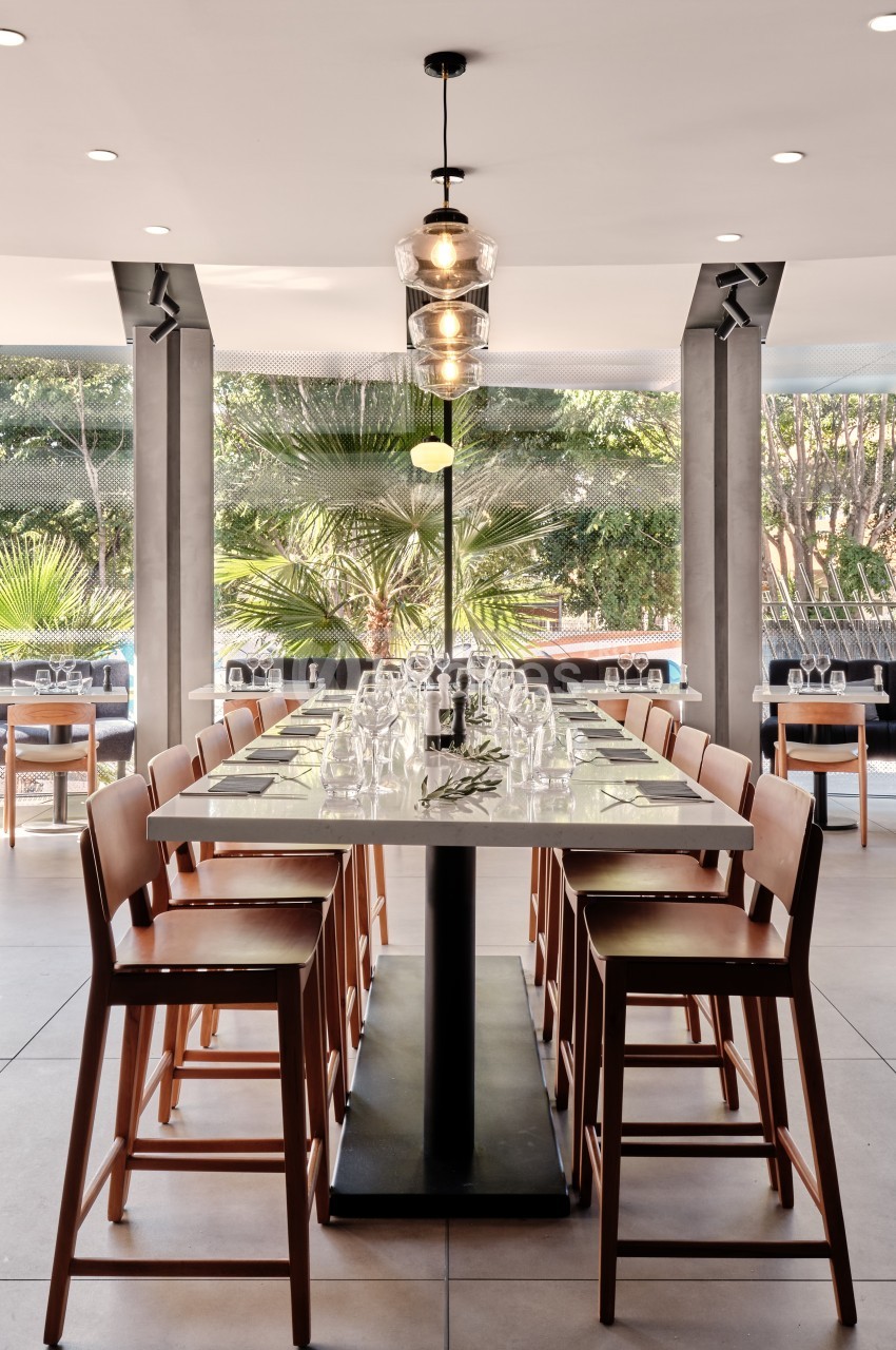 Table rectangulaire avec chaises en bois, dressée avec verres et couverts, dans un espace lumineux avec vue sur des palmiers.