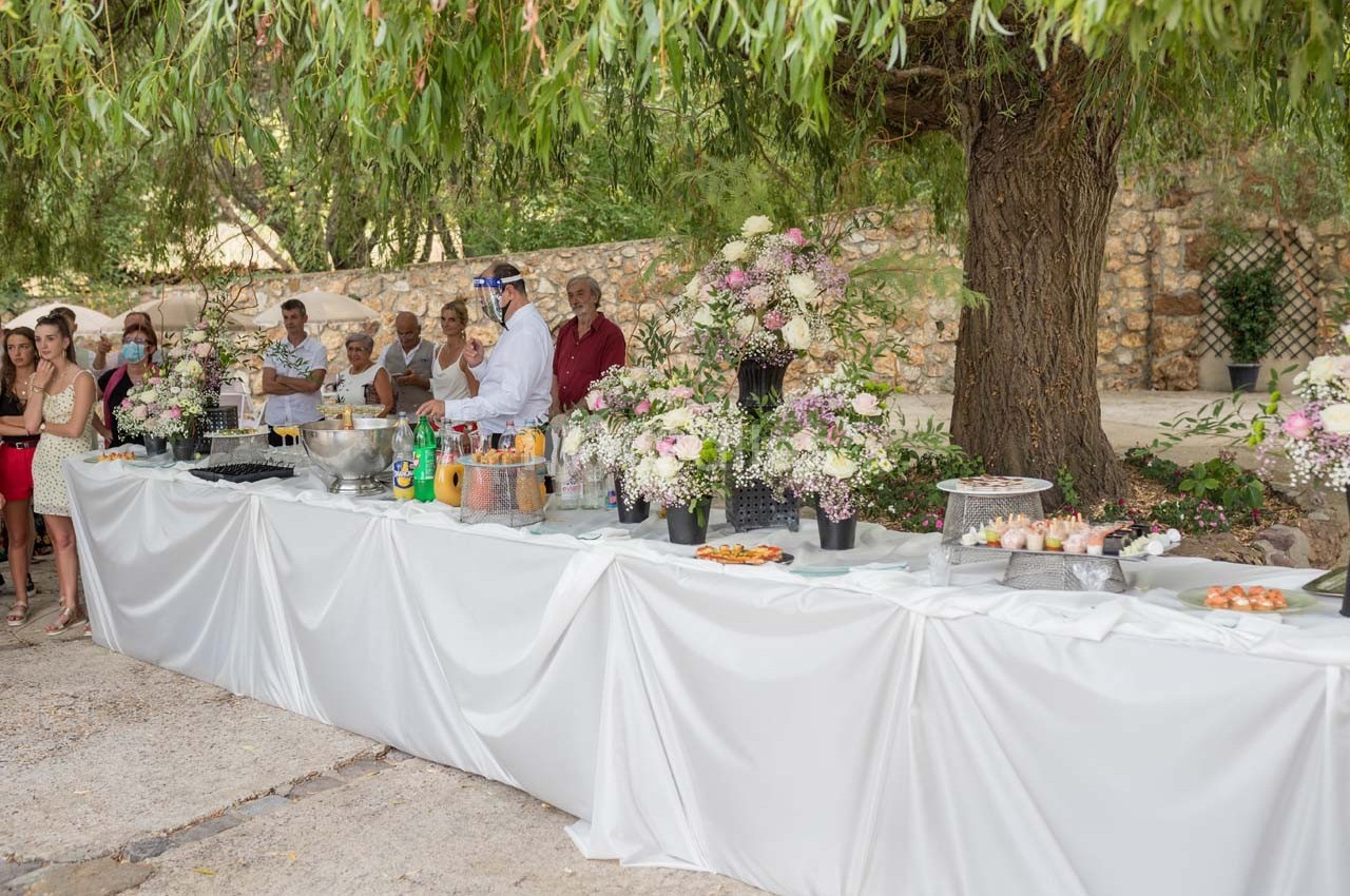 Buffet en plein air sous des arbres, avec des plats variés, des fleurs décoratives et des convives en arrière-plan.