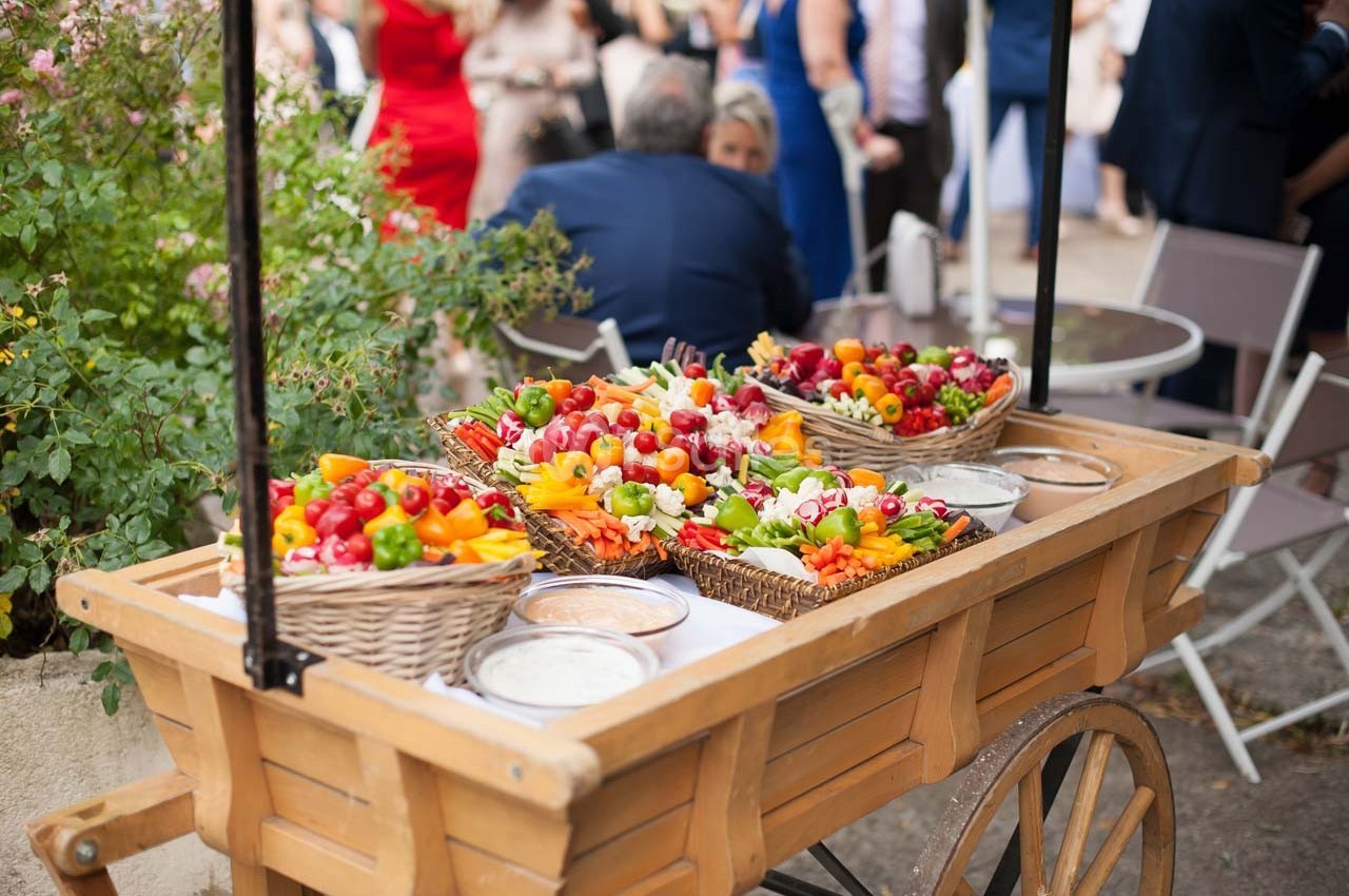 Chariot en bois rempli de paniers de légumes colorés avec des sauces, dans un cadre extérieur avec des invités en arrière…