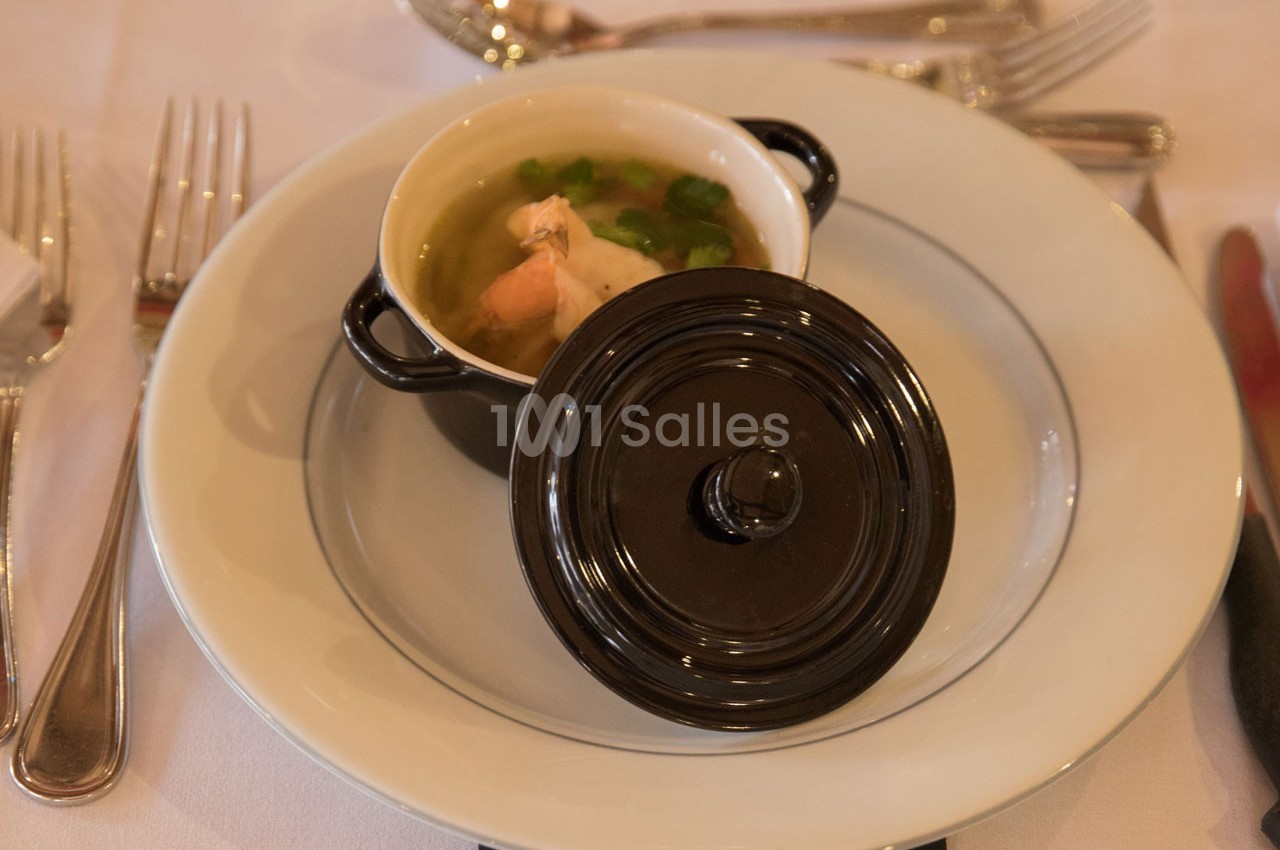 Petite cocotte noire contenant une soupe avec des légumes et un morceau de viande, présentée sur une assiette blanche.