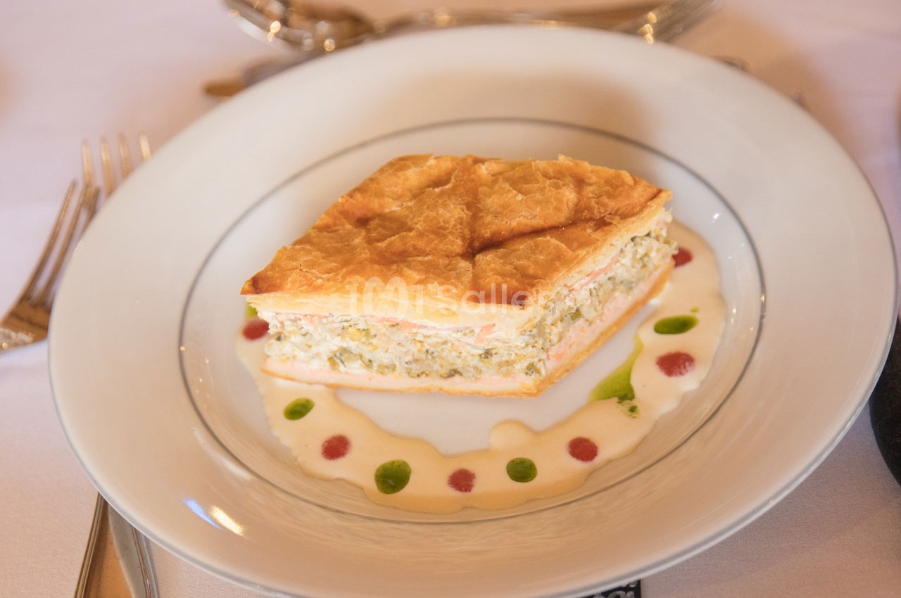 Part de feuilleté doré garni de farce crémeuse, servie sur une assiette avec une sauce décorative colorée.