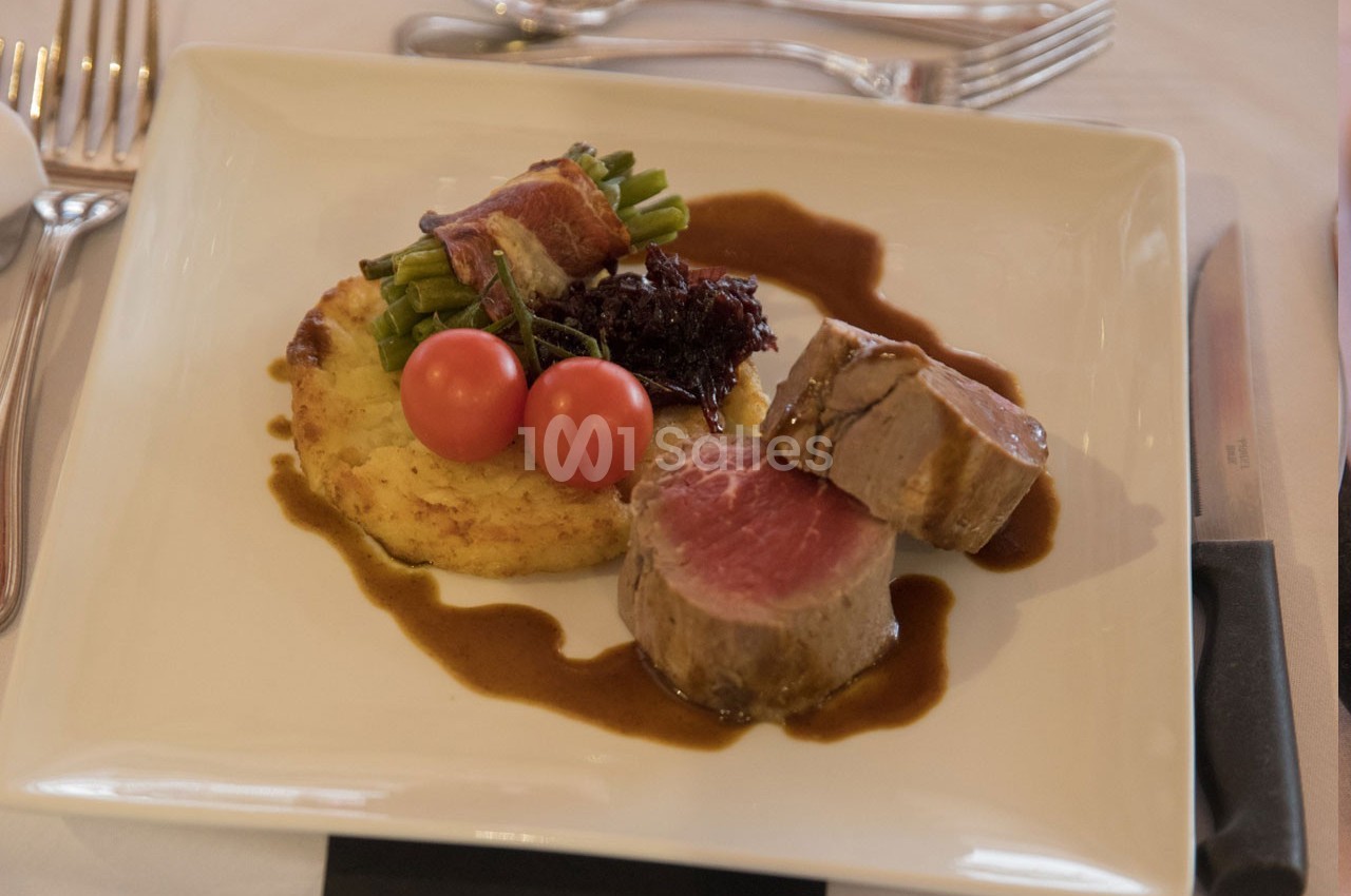 Assiette contenant un filet de viande, une galette de pommes de terre, des haricots verts enroulés de lard et des tomates…