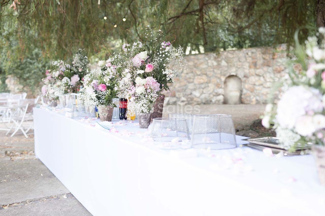 Table décorée pour un événement en extérieur, ornée de bouquets de fleurs roses et blanches sous des arbres.