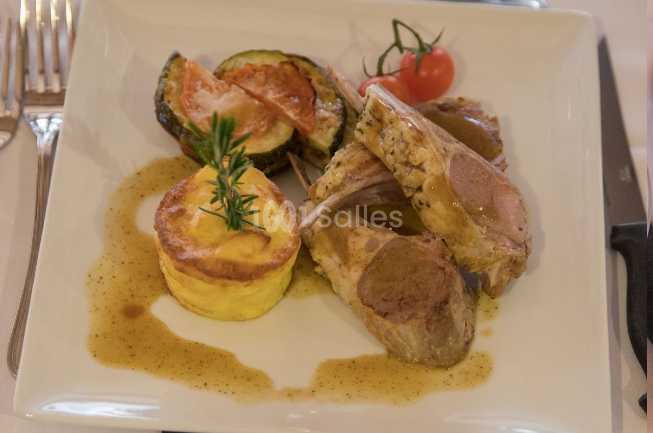 Assiette contenant des côtelettes d'agneau, un gratin individuel, des légumes grillés et une tomate cerise.