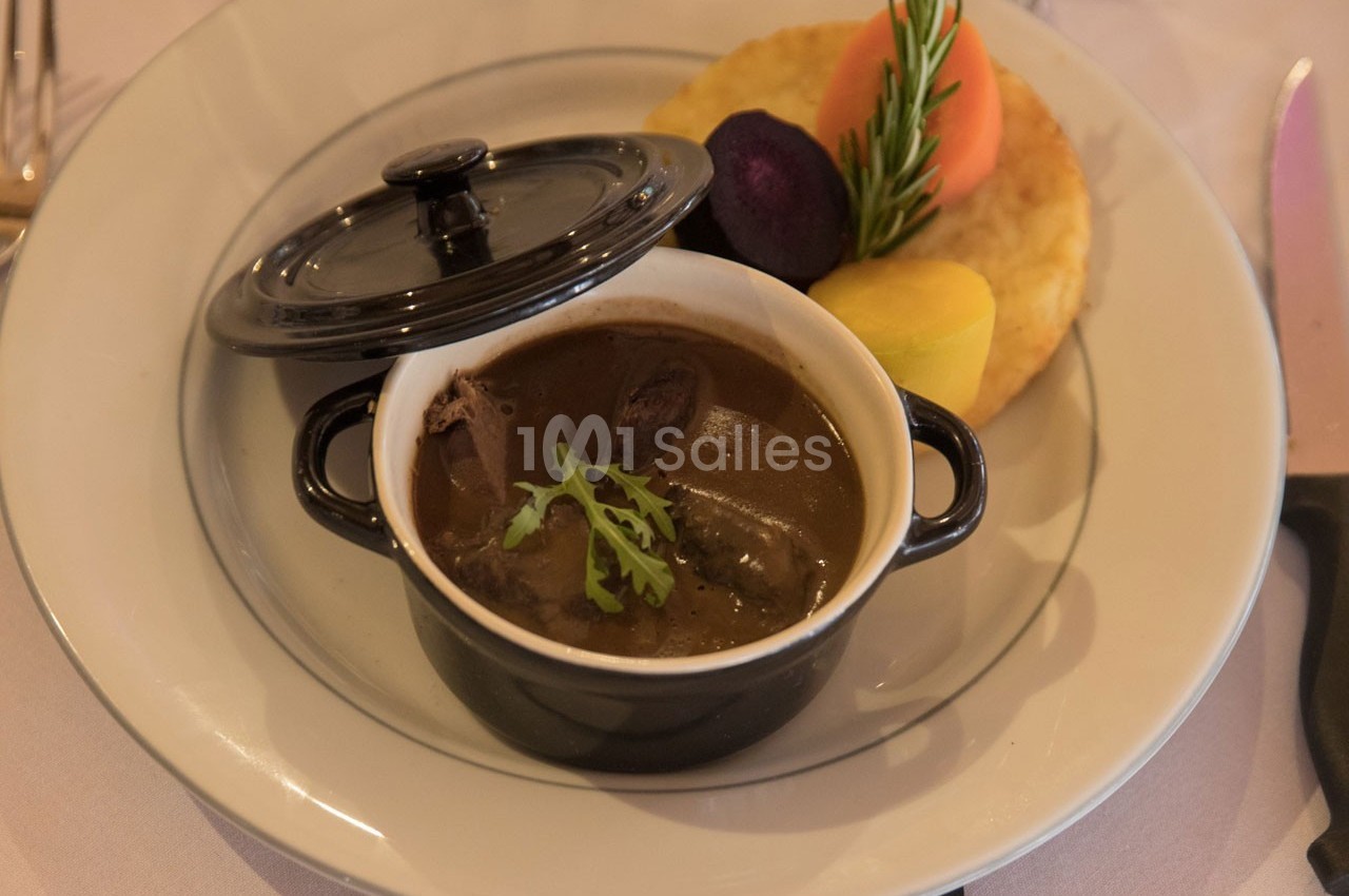 Plat de viande en sauce servi dans une cocotte noire, accompagné de légumes colorés et d'une galette dorée.