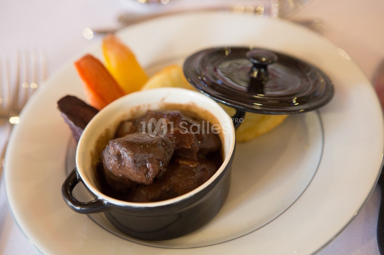 Plat de viande en sauce servi dans une petite cocotte noire, accompagné de légumes colorés sur une assiette blanche.