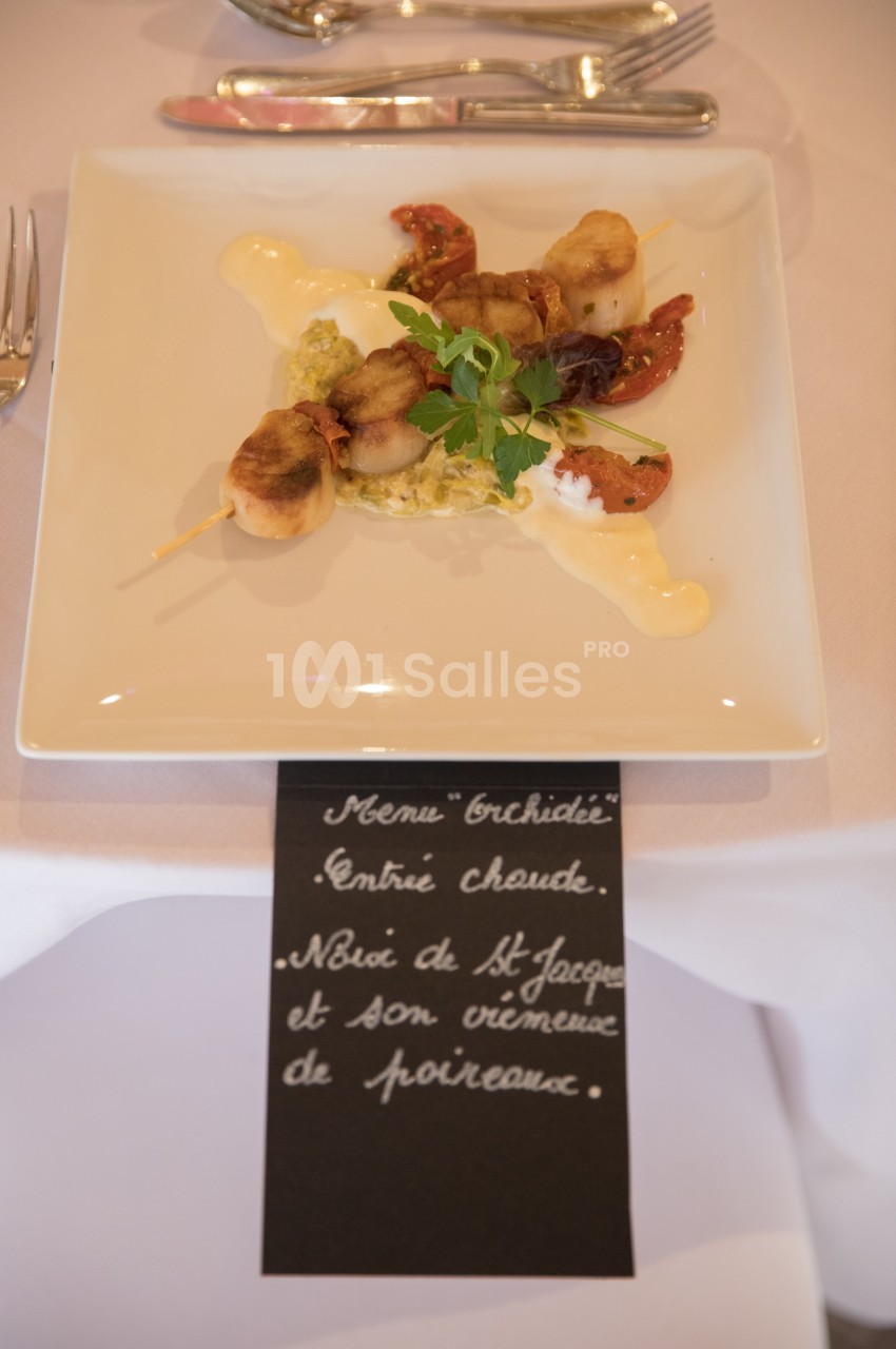 Brochette de noix de Saint-Jacques servie avec légumes, sauce crémeuse et décoration d'herbes sur une assiette carrée.