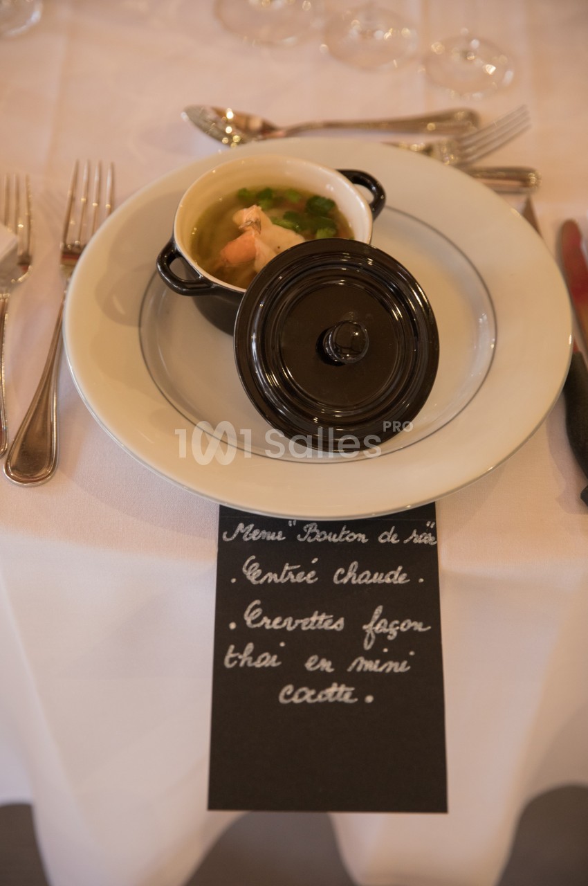 Mini cocotte contenant une entrée chaude aux crevettes, servie sur une table dressée avec couverts et ardoise descriptive.