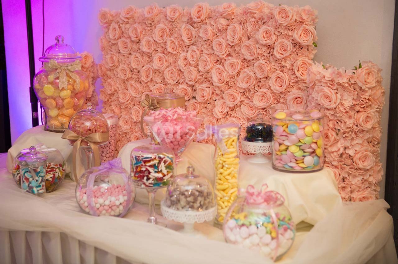 Table décorative avec des bonbonnières remplies de sucreries colorées, entourées de roses roses en arrière-plan.