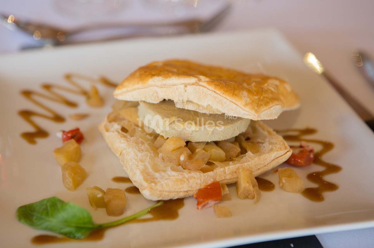 Feuilleté garni de foie gras et compotée de pommes, accompagné de sauce décorative et de légumes sur une assiette blanche.