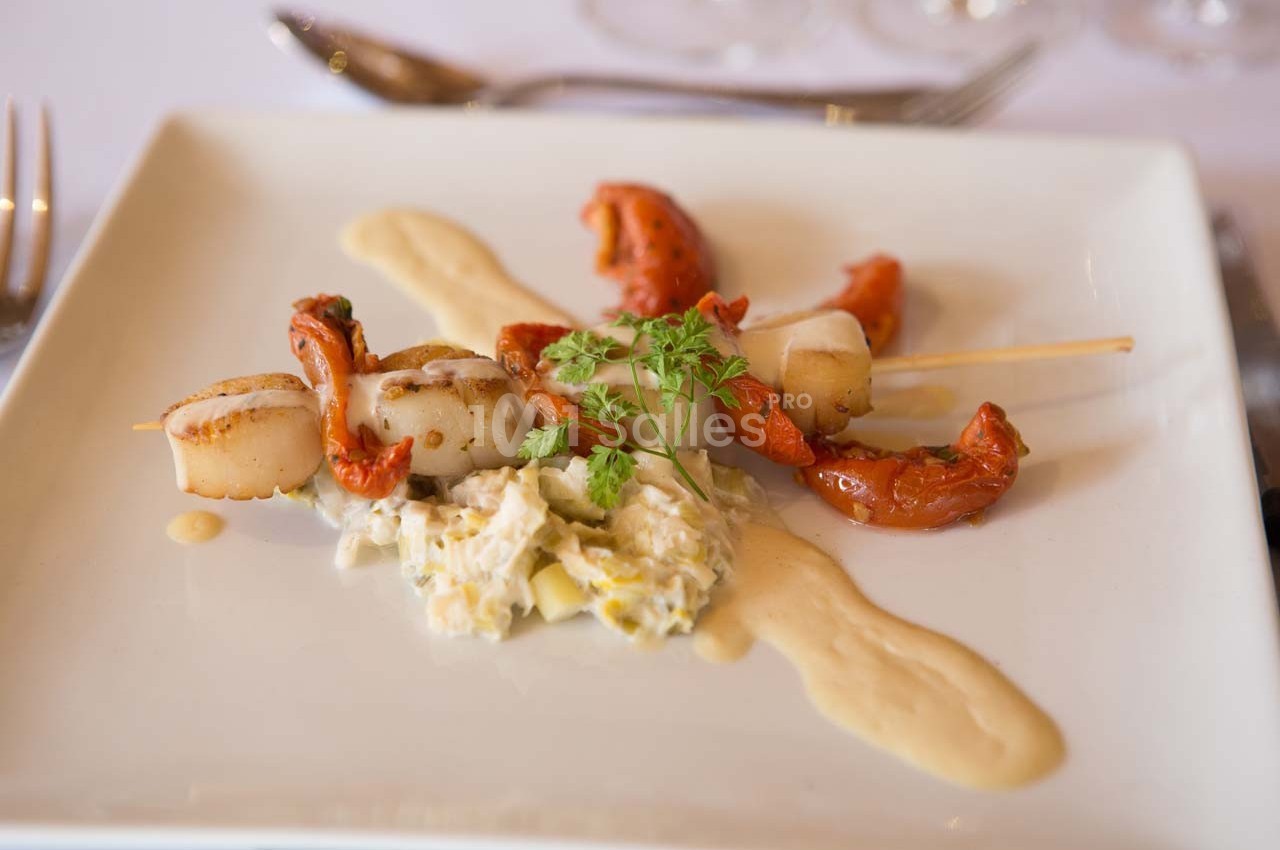 Brochette de noix de Saint-Jacques avec tomates confites, purée et salade crémeuse, servie sur une assiette blanche.