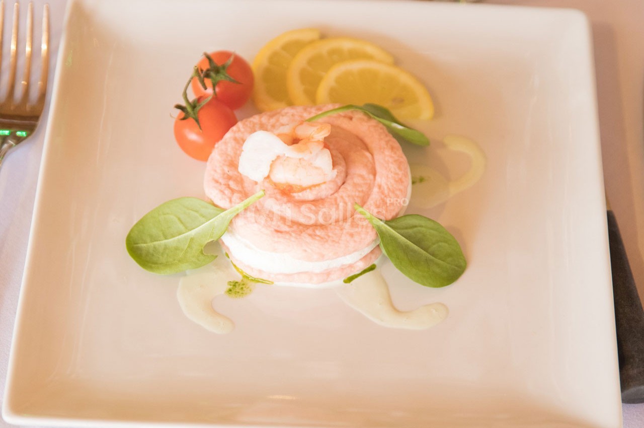 Entrée servie sur une assiette carrée avec mousse rose, crevette, feuilles de mâche, tomates cerises et tranches de citron.