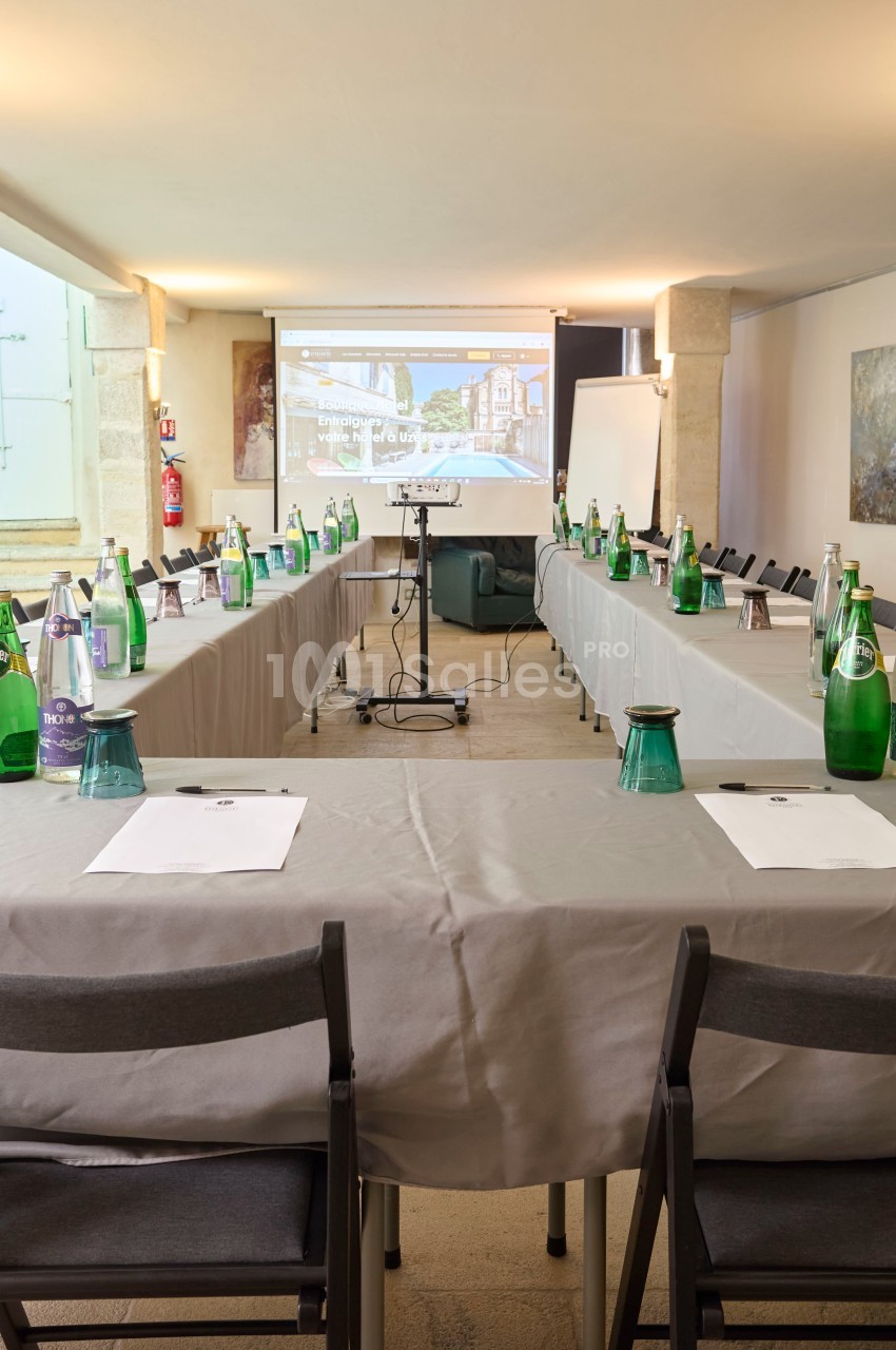 Salle de réunion aménagée en U avec tables couvertes, chaises, bouteilles d'eau, documents et écran de projection.