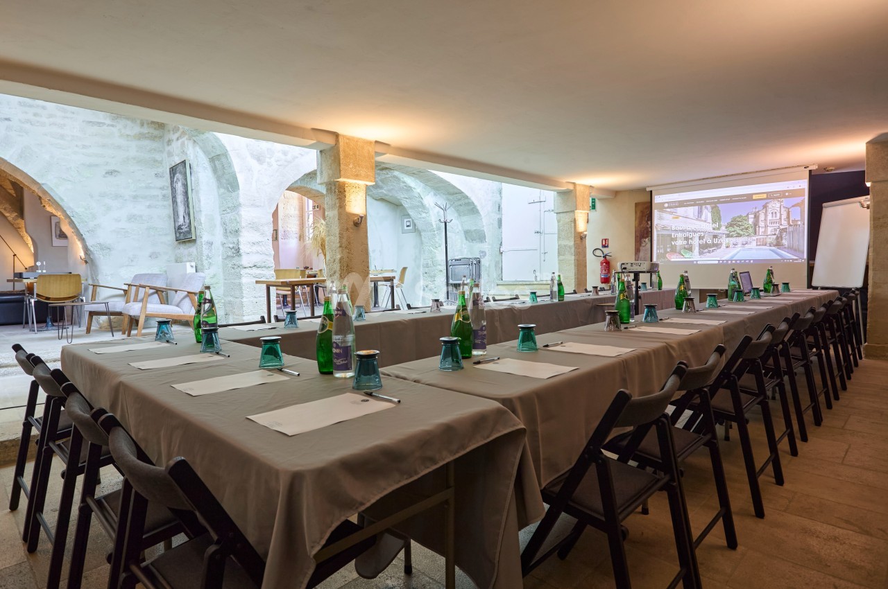 Salle de réunion avec tables disposées en U, chaises noires, bouteilles d'eau et écran de projection allumé.