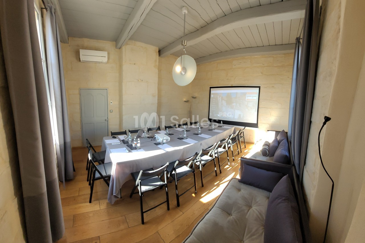 Salle de réunion lumineuse avec une grande table rectangulaire, chaises noires, canapé et écran de projection.