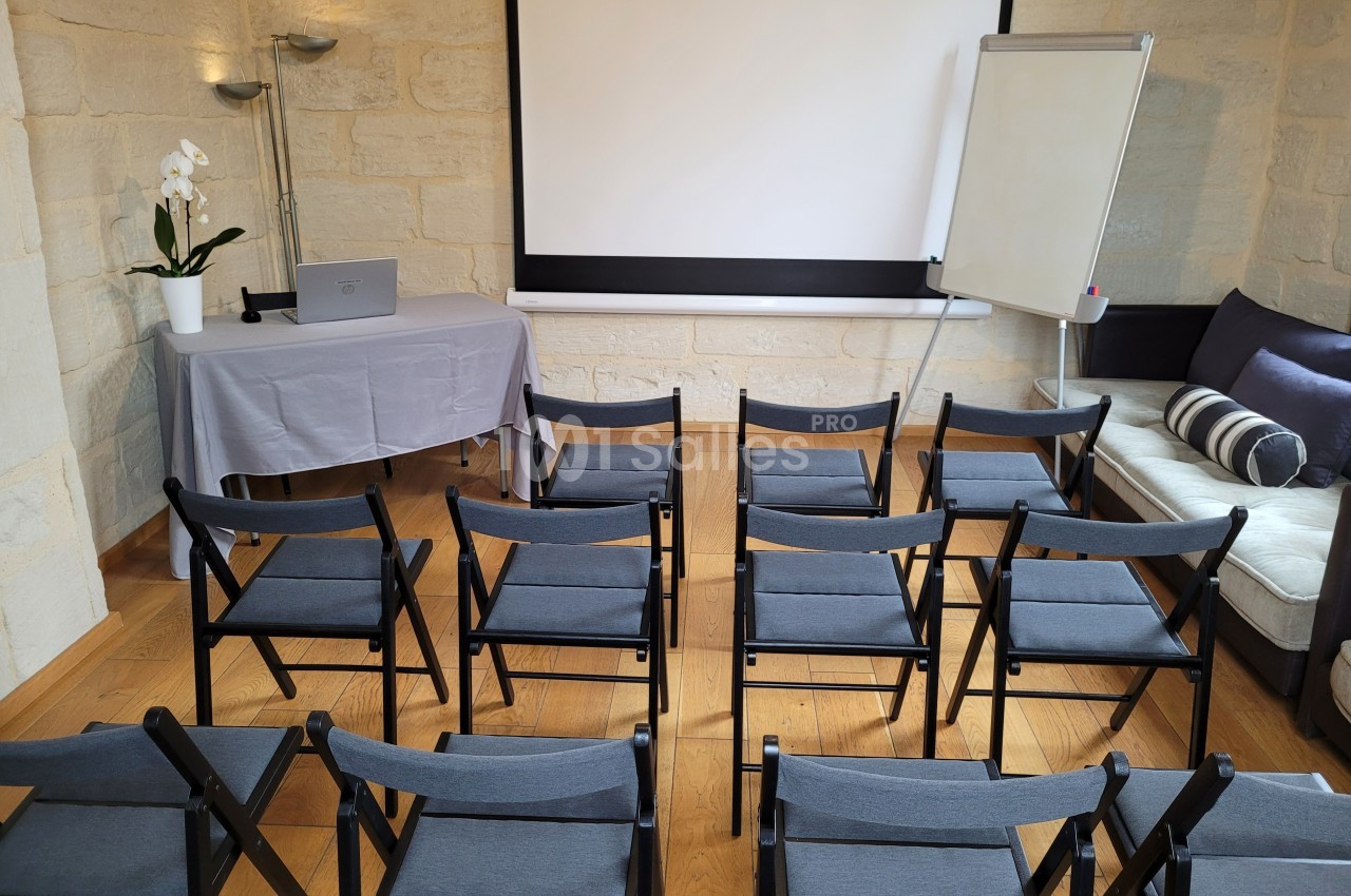 Salle de réunion équipée de chaises pliantes, d'un écran de projection, d'un tableau blanc et d'une table avec ordinateur.