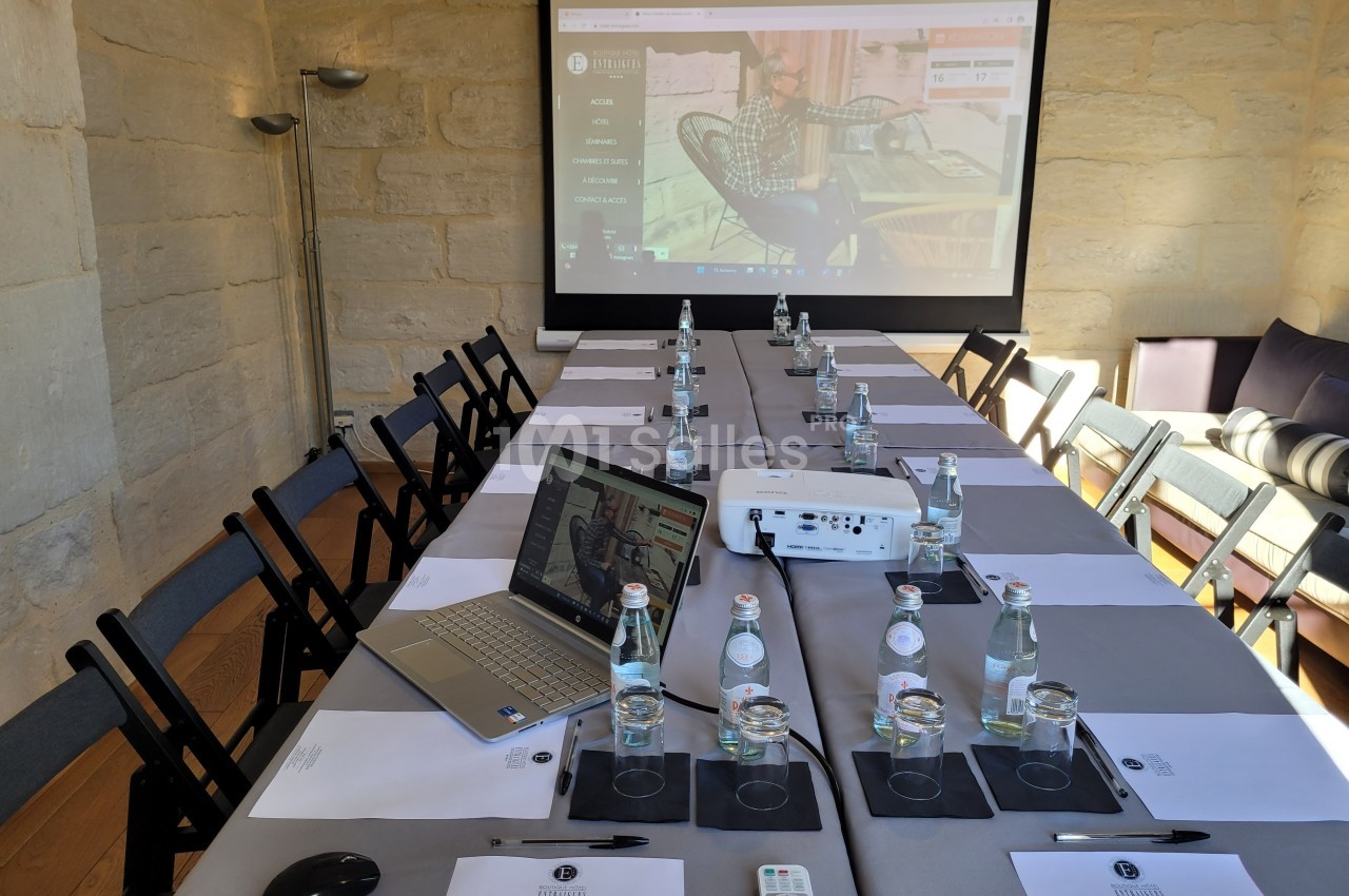 Salle de réunion équipée d'un projecteur, d'un écran, de chaises, de tables dressées avec bouteilles d'eau et documents.