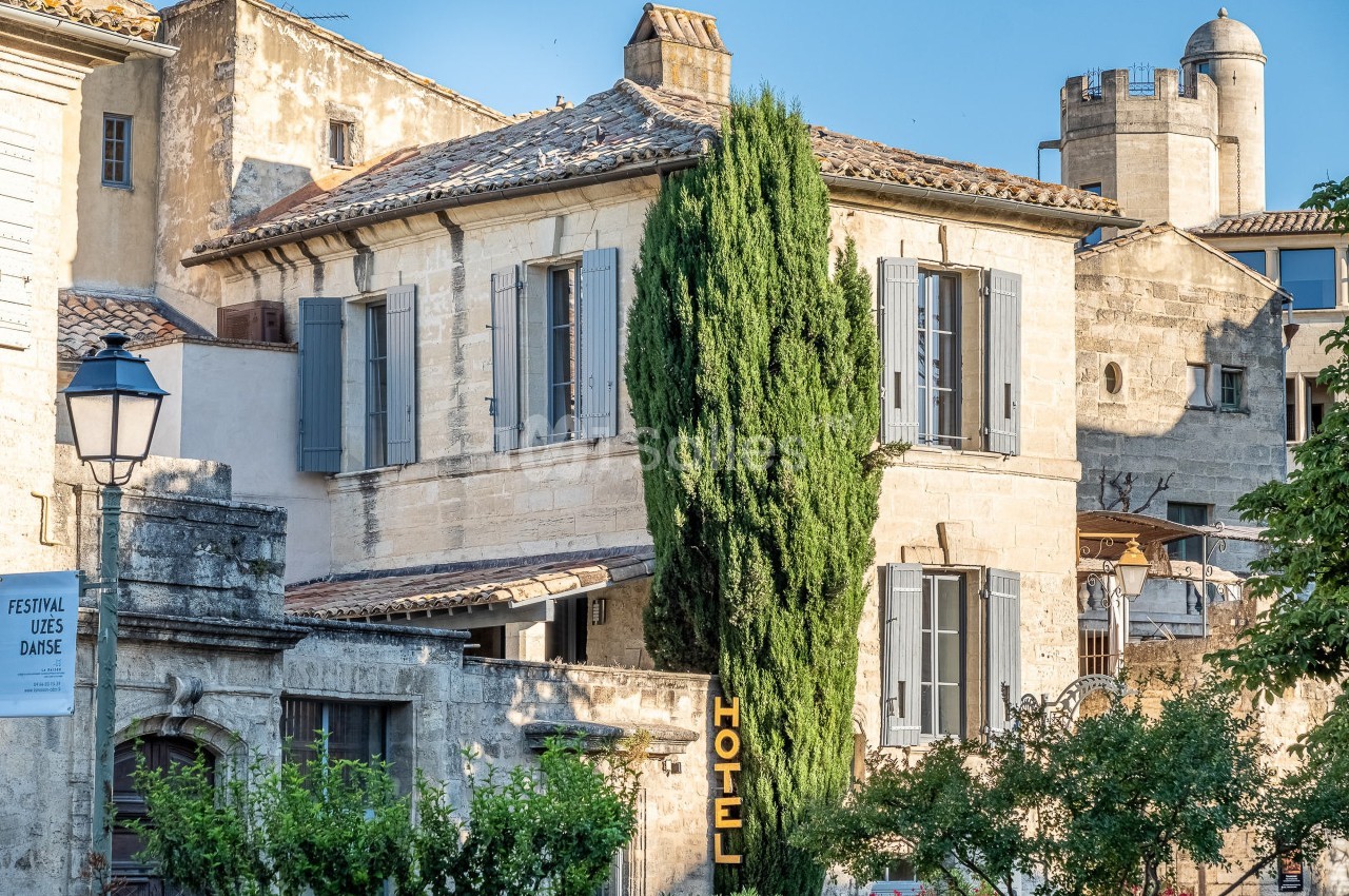 Façade en pierre d'un bâtiment ancien avec volets bleus, un cyprès et une enseigne d'hôtel dans un village historique.