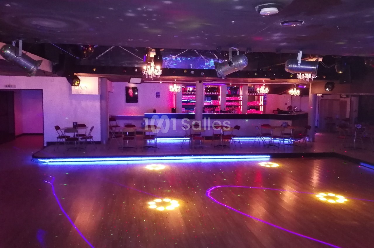 Salle de danse avec piste éclairée par des lumières colorées, bar illuminé en arrière-plan et chaises disposées autour.