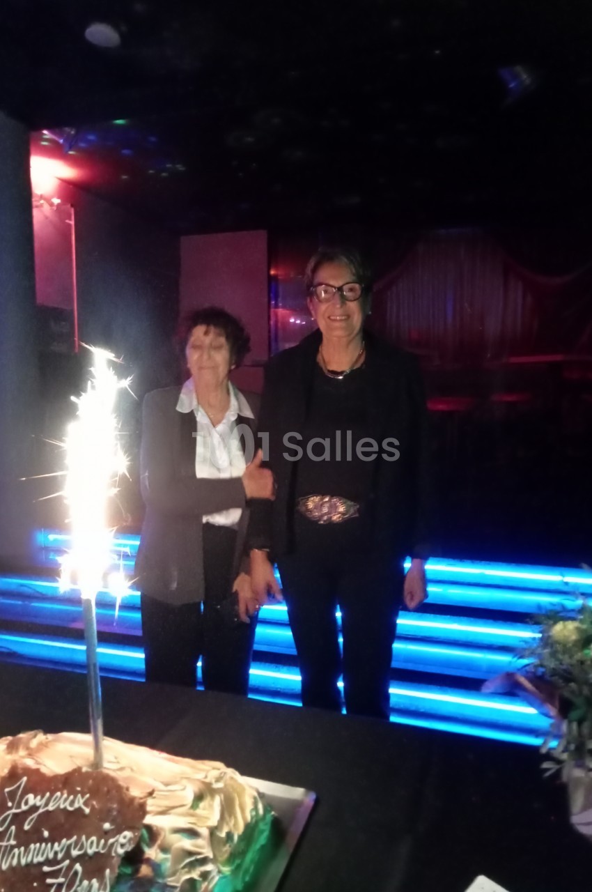 Deux femmes debout dans un environnement sombre près d'un gâteau d'anniversaire avec une bougie scintillante.