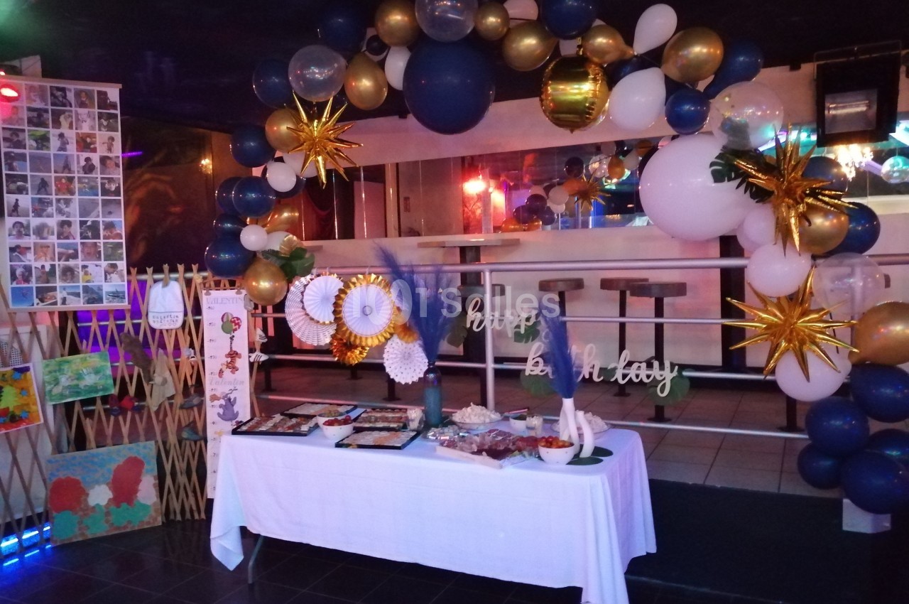 Décoration festive avec ballons bleus, blancs et dorés, table de buffet et panneaux décoratifs dans une salle sombre.