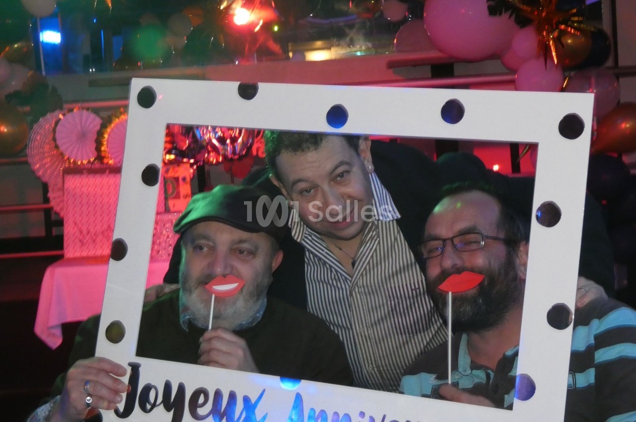 Trois hommes posent avec un cadre ’Joyeux Anniversaire’ et des accessoires amusants dans une ambiance festive.