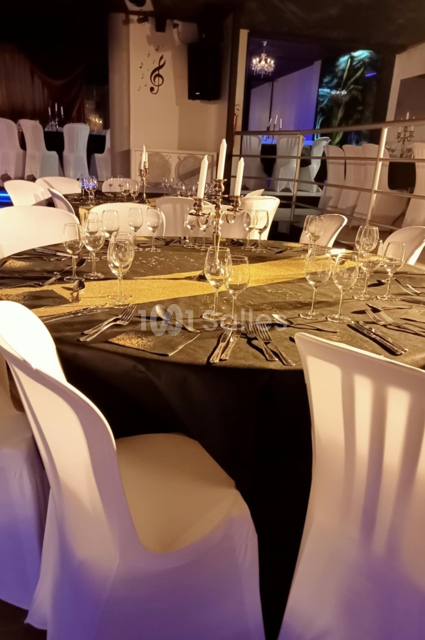 Table ronde élégamment dressée avec nappe noire, chemin de table doré, chandeliers et verres, entourée de chaises blanches.
