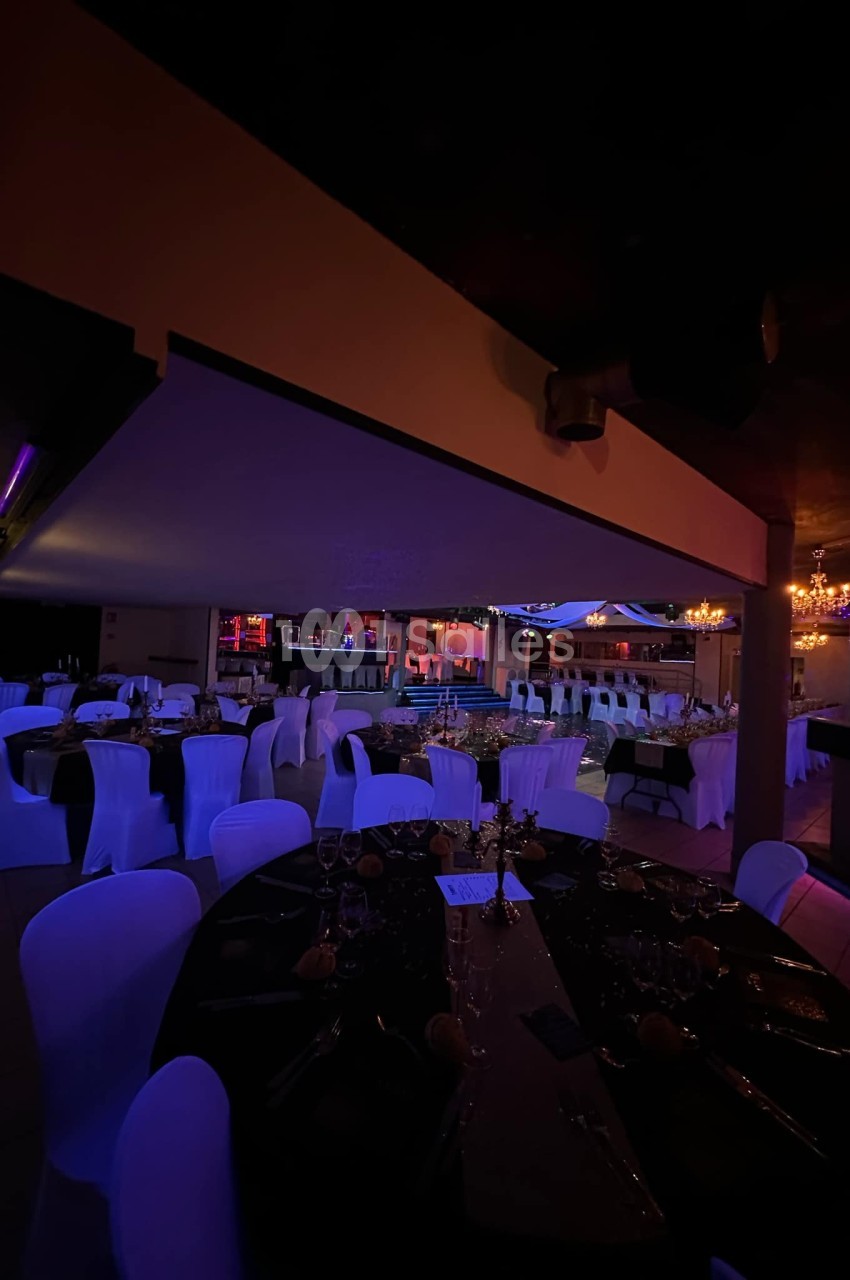 Salle de réception décorée avec des tables rondes, nappes noires, chaises blanches et éclairage tamisé.