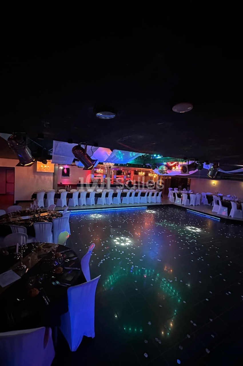Salle de réception sombre avec piste de danse illuminée, tables dressées et bar éclairé en arrière-plan.