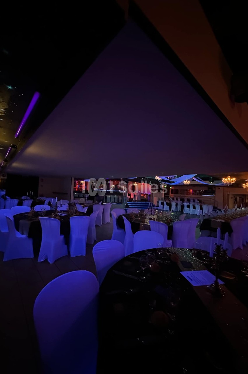 Salle de réception faiblement éclairée, avec des tables dressées et des chaises couvertes de housses blanches.