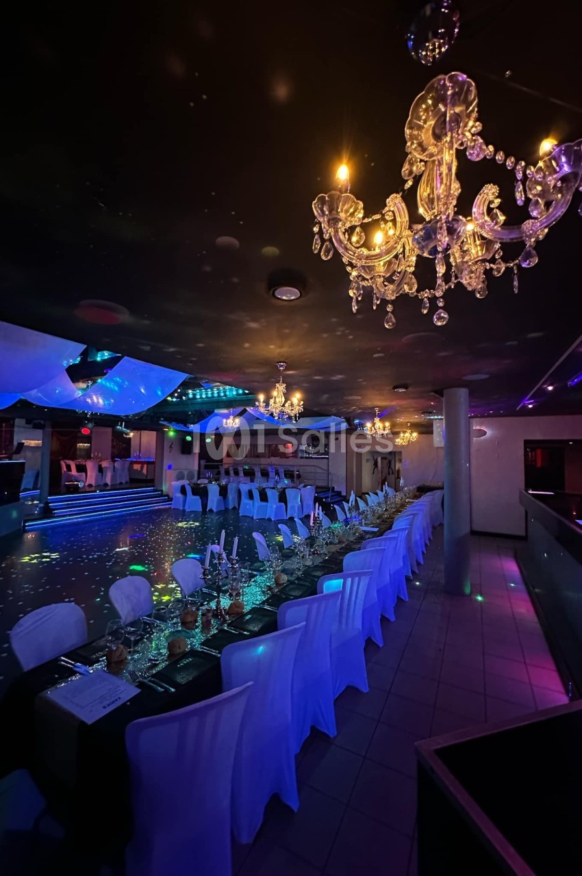 Salle de réception décorée avec des tables alignées, nappes blanches, chandeliers suspendus et éclairage tamisé coloré.