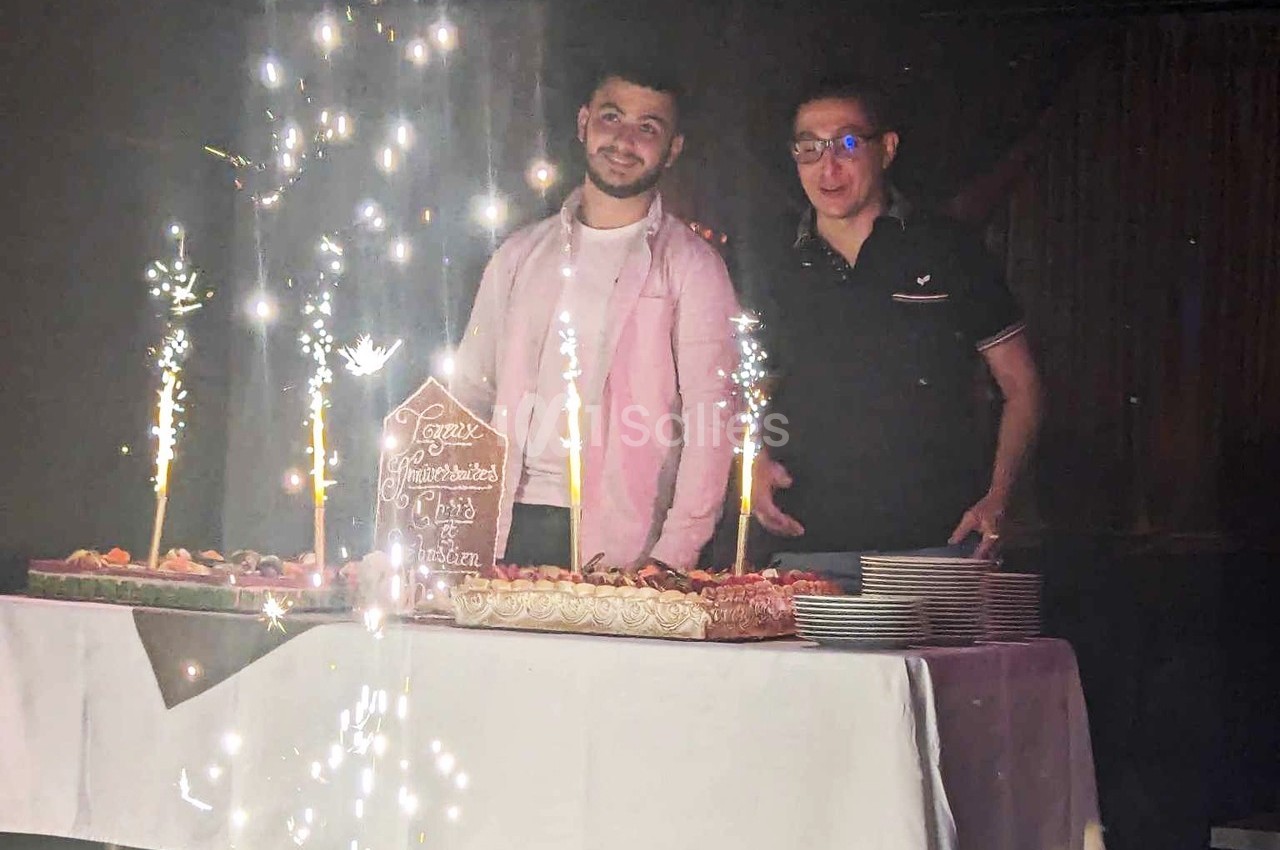 Deux hommes debout derrière une table avec des gâteaux décorés et des cierges étincelants allumés.