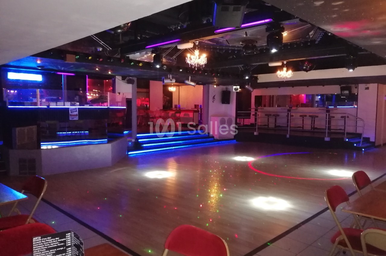 Salle de danse vide avec piste en parquet, éclairages colorés, tables et chaises autour, ambiance de soirée.