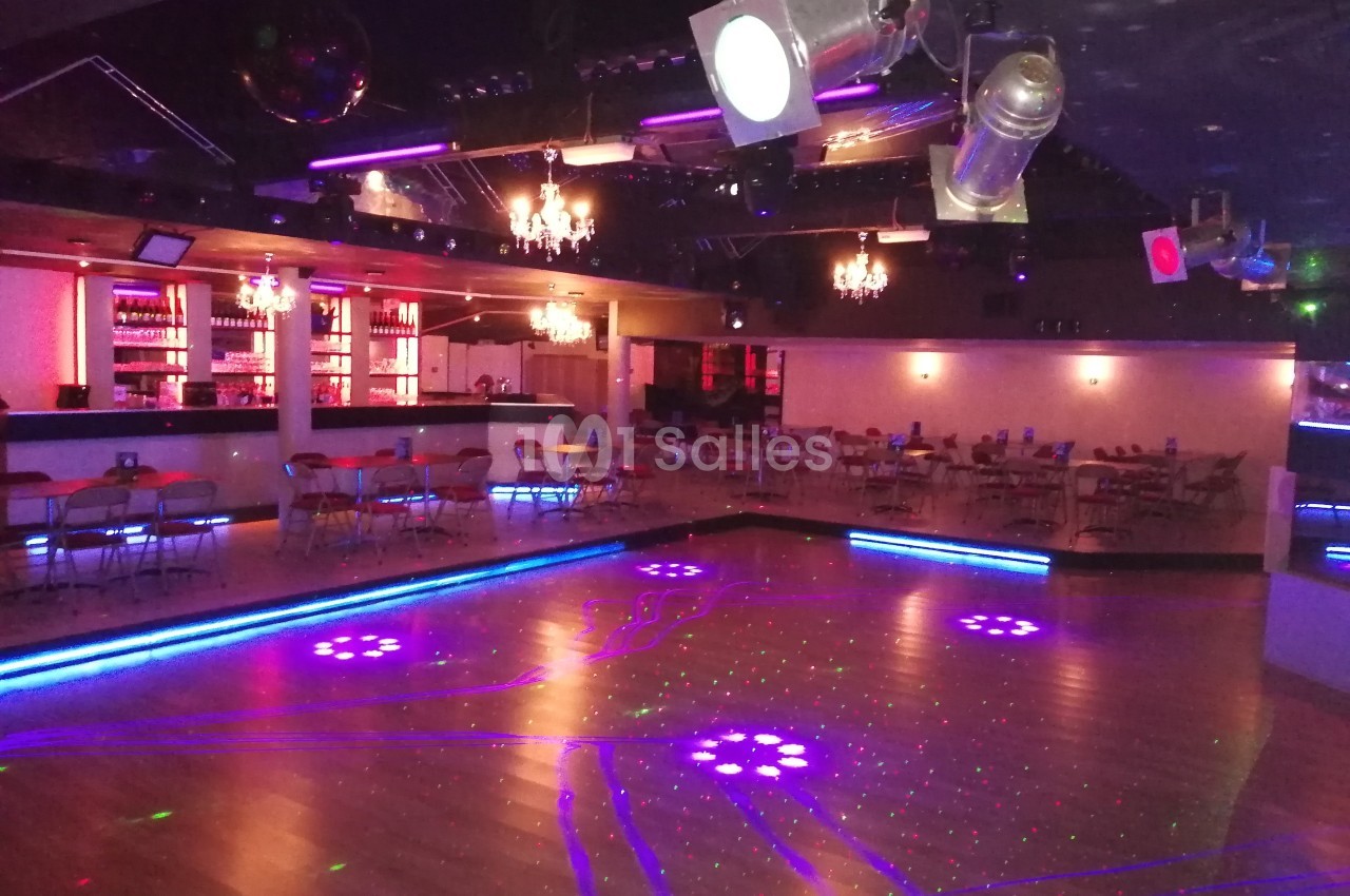 Salle de danse avec éclairage coloré, piste en bois, tables et chaises disposées près d'un bar illuminé.